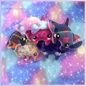 May include: A collection of five plush stingray toys featuring various patterns and colors, including pink galaxy, dark plaid, bright pink, purple, and a multi-colored confetti print. Each plush has embroidered eyes and the text underwatercircus.