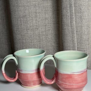 May include: Two ceramic mugs featuring a two-tone design with a pale mint green upper section and a dusty rose pink lower section. Each mug has a curved handle and a slightly flared rim, showcasing a smooth, glossy finish with visible horizontal texture.