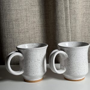 May include: Two ceramic mugs with a white speckled glaze and a smooth finish. Each mug features a wide, rounded handle and a flared rim. The base of each mug is unglazed, revealing a natural tan clay color. The mugs have a cylindrical body.