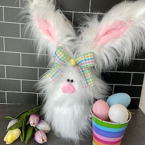 May include: A fluffy white Easter bunny decoration with large ears lined in pink. It has a pink nose, black eyes, and a plaid bow. A small rainbow bucket holds speckled eggs. Tulips in shades of purple and yellow are also present.