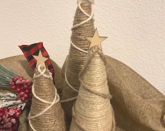 May include: Three cone-shaped decorative trees wrapped in tan twine, each topped with a small wooden star. The trees are accented with thin white or brown string spiraling around them. A red and black plaid ribbon and red berries are visible.