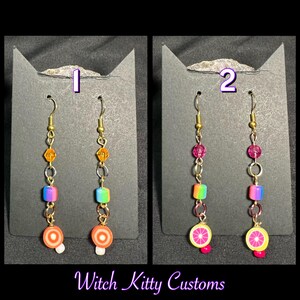 Op de afbeelding: Twee paar handgemaakte oorbellen. Eén paar heeft oranje, regenboog en witte elementen, met een cirkelvormig ontwerp aan de onderkant. Het andere paar heeft roze, regenboog en gele elementen, met een citrusvruchtenontwerp. De tekst "Witch Kitty Customs" staat onderaan.