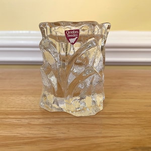 May include: A clear glass votive candle holder with a textured, carved design. The candle holder has a thick base and a flared top. The Orrefors Sweden logo is visible on the front. The candle holder is sitting on a wooden surface.