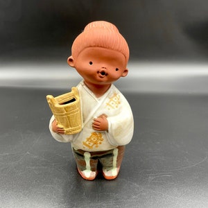 May include: A small ceramic figurine of a person with brown skin, wearing a white robe with gold accents and holding a small brown bucket. The figurine has a brown bun on its head and is smiling.