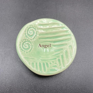 May include: A round, light green ceramic dish with embossed spiral and linear designs. The word "Angel" is printed in brown. The dish has a slightly irregular edge and is set against a dark background.