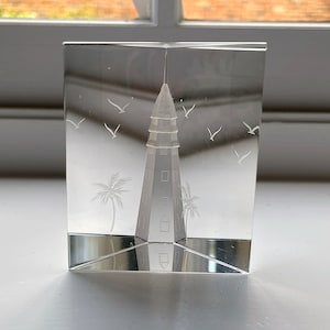 May include: A clear glass paperweight with an etched design of a lighthouse, palm trees, and birds in flight.