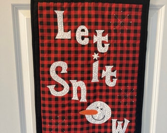 May include: A red and black buffalo plaid fabric banner with white lettering that reads "Let It Snow". A white snowman with a black smile and orange nose is in the lower right corner of the banner.