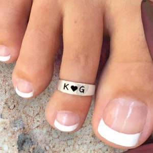 May include: A silver-toned metal toe ring featuring the engraved letters K and G separated by a small black heart symbol. The band is smooth and polished, worn on the second toe of a foot with a French manicure.