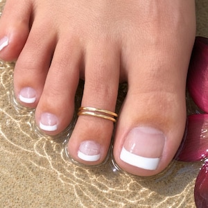 May include: A gold-toned metal toe ring featuring a double-band design worn on the second toe. The toes have a French manicure with white tips. A dark pink flower petal is visible on the right side, and the toes are partially submerged in water.