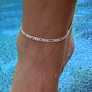 May include: A silver chain anklet with a delicate, figaro design. The anklet is worn on a person's ankle, with the foot resting on a textured surface.
