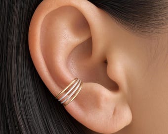 May include: A gold-toned ear cuff featuring a multi-band design with three parallel, thin, curved wires that wrap around the outer edge of the ear cartilage. The piece has a polished metallic finish and is positioned on the middle of the ear.