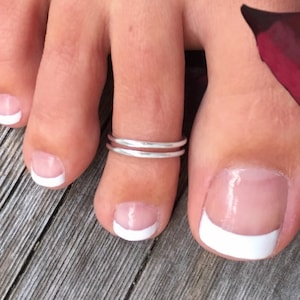 May include: A silver toe ring with a double band design. The ring is worn on the second toe of a foot with white nail polish.