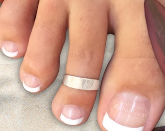 May include: A smooth, polished silver-toned metal toe ring worn on the second toe. The toe ring features a simple, solid band design with a reflective surface. The foot displays a classic French manicure with white tips on the toenails.
