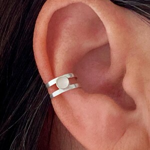 May include: A silver ear cuff with a circular, light-colored stone in the center. The cuff has a double-band design and is worn on the ear. The image shows a close-up of the ear and the jewelry.