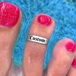 May include: A silver toe ring with the word "Custom" engraved on it. The ring is worn on a toe with pink nail polish. The toe is in a pool of blue water.