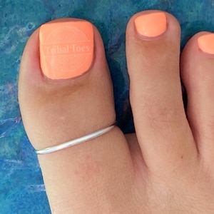May include: Close-up of a foot with orange nail polish and a silver toe ring. The big toe has the text "Tribal toes" printed on it. The background is a textured blue surface.