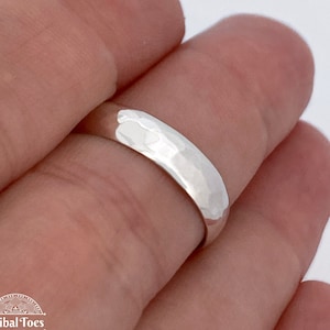 May include: A silver ring with a hammered texture. The ring is smooth on the inside and has a slightly rounded shape. The ring is held between two fingers.