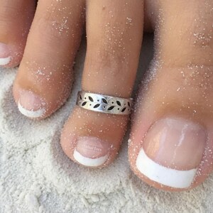 May include: A silver-toned metal toe ring featuring a repeating pattern of black-filled leaf shapes. The band has a smooth, polished finish and is worn on the second toe, which has a white-tipped French manicure and is covered in fine sand.