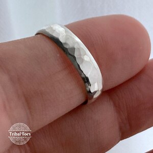 May include: A hammered silver ring with a textured surface. The ring is round and appears to be worn on a finger. The ring has a shiny, reflective quality. The logo "Tribal Toes" is visible in the lower left corner.