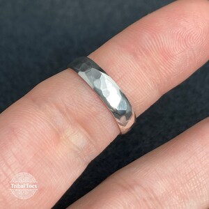 May include: A silver ring with a hammered texture. The ring is shown on a finger, with a dark background. The ring has a faceted surface, reflecting light. The ring is a simple band design, suitable for everyday wear.