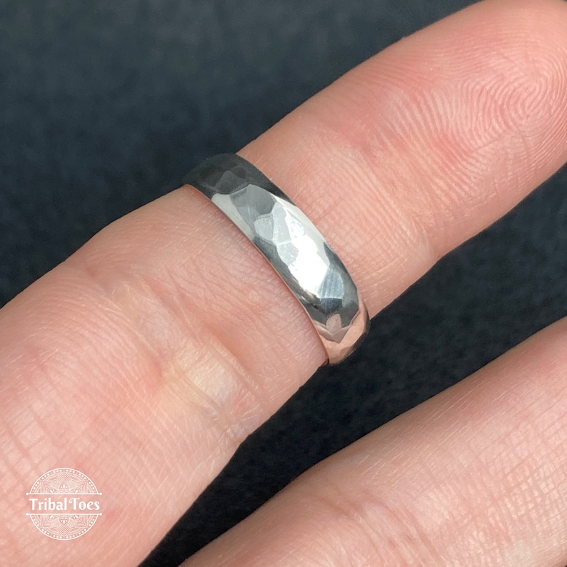 May include: A silver ring with a hammered texture. The ring is shown on a finger, with a dark background. The ring has a faceted surface, reflecting light. The ring is a simple band design, suitable for everyday wear.
