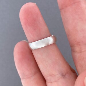 May include: A silver ring with a brushed finish is shown on a finger. The ring has a simple, classic design with a smooth, rounded band. The ring is likely made of sterling silver or a similar metal.