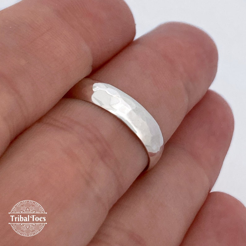 May include: A silver ring with a hammered texture. The ring is smooth on the inside and has a slightly rounded shape. The ring is held between two fingers.
