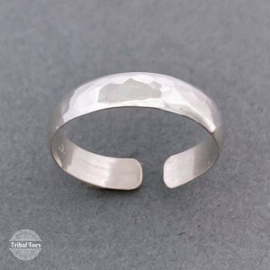 May include: A silver toe ring with a hammered texture. The open design allows for an adjustable fit. The ring is displayed on a gray surface. The Tribal Toes logo is visible in the bottom left corner.