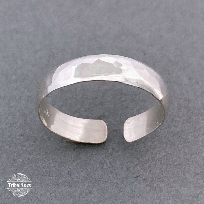 May include: A silver toe ring with a hammered texture. The open design allows for an adjustable fit. The ring is displayed on a gray surface. The Tribal Toes logo is visible in the bottom left corner.