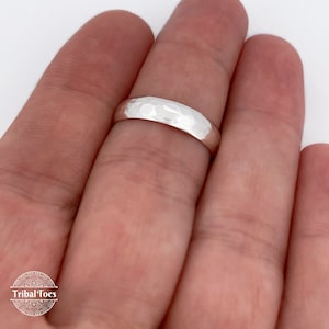 May include: A hammered silver ring with a textured surface. The ring is a simple band design, reflecting light. The ring is shown on a finger against a white background. The ring is a jewelry item.