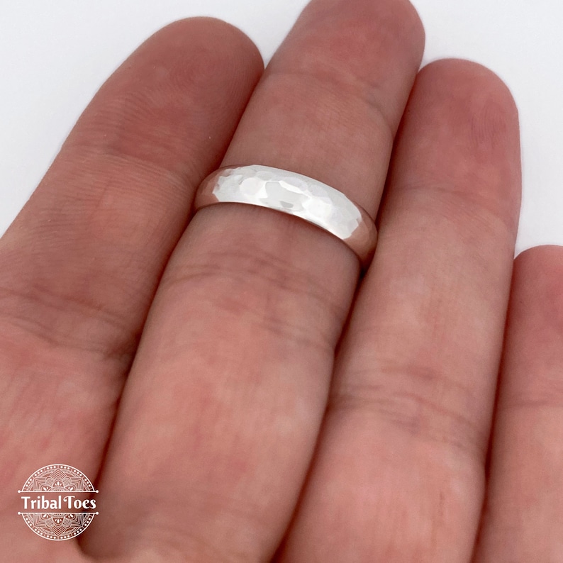 May include: A hammered silver ring with a textured surface. The ring is a simple band design, reflecting light. The ring is shown on a finger against a white background. The ring is a jewelry item.