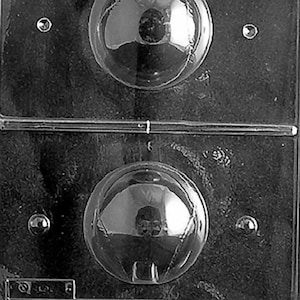 May include: A clear plastic mold with two round cavities for making chocolate ornaments. The mold is designed to create a sphere shape with a small indentation at the top.