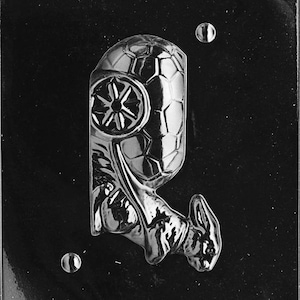 May include: A black plastic mold for making chocolates or candies. The mold features a detailed design of a character with a helmet and a star emblem. The mold is for a party or event.