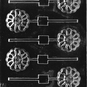 Puede incluir: Un molde de plástico transparente para hacer cinco caramelos en forma de piruleta. Cada caramelo tiene un diseño de sol con una cara sonriente. El molde tiene cinco palos para sujetar las piruletas.