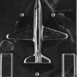 May include: A clear plastic airplane-shaped mold for making chocolates or candies. The mold features a detailed airplane design with wings, a fuselage, and a tail. The mold also has two rectangular cavities and three circular cavities.