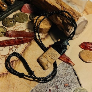 Puede incluir: Un collar de cordón negro con un colgante de piedra rectangular. La piedra es de color marrón claro con vetas marrones más oscuras. El colgante está atado al cordón con una cuerda negra.