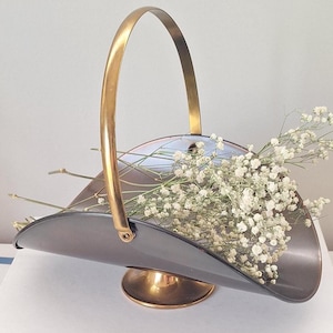 May include: A vintage metal basket with a gold handle and base, holding white flowers. The basket has a dark grey finish and is placed on a stack of white books, creating a simple, elegant display.