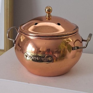 Peut inclure: Un pot-pourri en cuivre avec un couvercle et de petites poignées. Le pot a une forme ronde et une finition brillante. Le mot "POTPOURRI" est imprimé sur le devant du pot. Le pot est posé sur deux livres blancs.