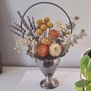 May include: A silver-toned metal pedestal vase with a ruffled rim and a circular handle, filled with a dried floral arrangement. The bouquet includes purple lavender, yellow billy balls, orange and cream strawflowers, and green eucalyptus leaves.