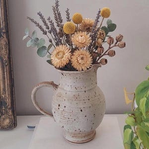 May include: A speckled cream ceramic pitcher filled with a dried floral arrangement. The bouquet includes yellow billy balls, lavender sprigs, pale strawflowers, eucalyptus leaves, and small brown seed pods, creating a textured, neutral-toned display.
