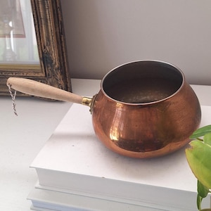 May include: A round copper pot with a polished metallic finish, featuring a single long wooden handle attached by a brass ferrule. The pot has a wide, open top and a rounded base, with a small piece of string tied to the end of the wooden handle.