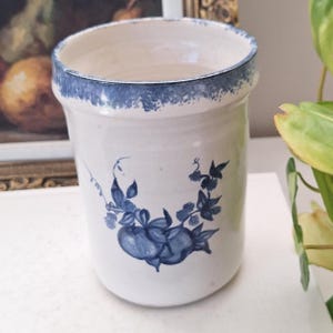 May include: A cylindrical ceramic utensil crock with a white base and a blue sponge-painted rim. The front features a hand-painted blue botanical illustration of two round fruits surrounded by leaves and curling vines on a smooth, glossy surface.