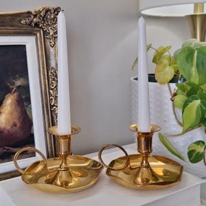 May include: A pair of polished gold-tone brass candle holders, each featuring a circular base with a scalloped edge, a small side handle, and a central socket holding a slender white taper candle. The metal surface is reflective and smooth.
