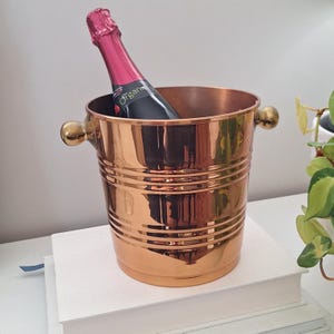 May include: A polished copper-coloured metal ice bucket with a ribbed exterior design and two rounded side handles. A bottle with a dark label and a bright pink foil-wrapped neck is placed inside the bucket, extending from the top opening.