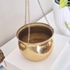 May include: A polished, gold-toned hanging plant pot with a rounded bowl shape. It is suspended by fine chains. The interior of the pot reflects the light. A decorative item for indoor plants.