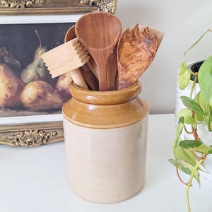 May include: A selection of wooden kitchen utensils, including spoons and a masher, are stored in a beige and brown ceramic jar. The jar is on a white surface, with a framed painting and a potted plant in the background.