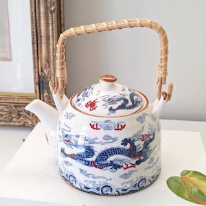 Peut inclure: Théière en céramique blanche avec un motif de dragon bleu et rouge. La théière a une anse tressée et un couvercle assorti. Le motif du dragon est répété sur le couvercle et le corps de la théière. C'est un objet décoratif.