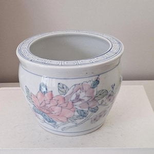 May include: A white ceramic planter with a rounded body and a wide opening. The exterior features a hand-painted floral design in soft pink and green tones, accented by a blue geometric Greek key pattern encircling the top rim of the vessel.
