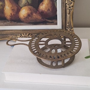 Peut inclure: Un dessous de plat circulaire en laiton présentant un motif ajouré complexe avec une série de petites découpes géométriques répétitives le long du bord extérieur et un anneau central. Le support en métal inclut une petite poignée incurvée.