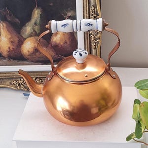 May include: A polished copper tea kettle with a rounded body and a curved spout. The handle and the lid knob are made of white ceramic, featuring delicate blue painted floral and cross-hatch patterns. The kettle has a metallic finish.